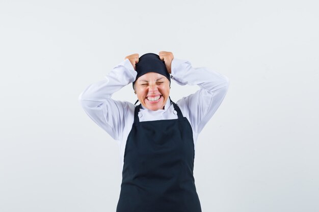 Cocinera cogidos de la mano en la cabeza en uniforme, delantal y mirando feliz, vista frontal.