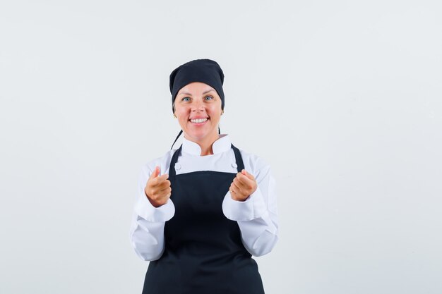 Cocinera fingiendo sostener algo en uniforme, delantal y luciendo optimista. vista frontal.