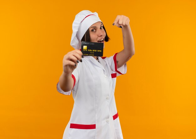 Cocinera joven alegre en uniforme de chef estirando la tarjeta de crédito y apuntando a ella aislada en naranja