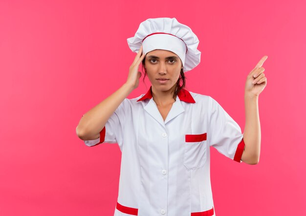 Cocinera joven cansada con uniforme de chef apunta al lado poniendo la mano en la cabeza con espacio de copia