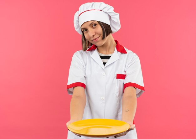 Cocinera joven complacida en uniforme de chef estirando la placa hacia el frente aislado en la pared rosa