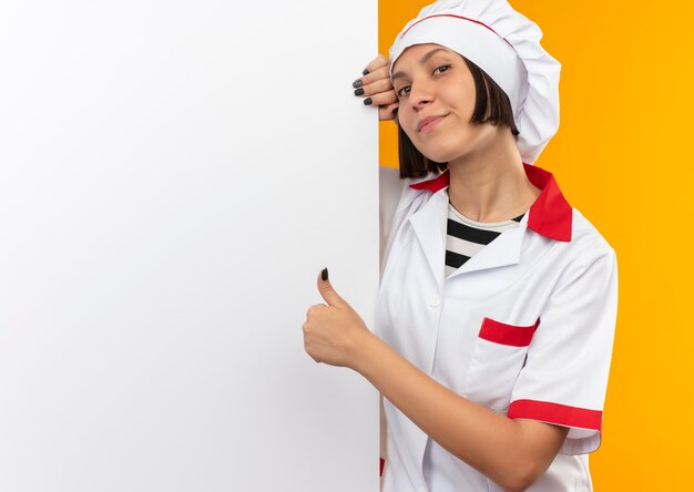 Cocinera joven complacida en uniforme de chef de pie detrás de una pared blanca y mostrando el pulgar hacia arriba aislado en la pared