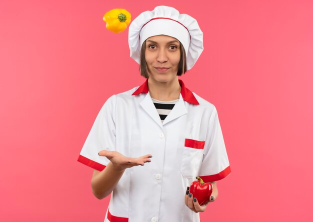 Cocinera joven complacida en uniforme de chef sosteniendo y arrojando pimientos aislados sobre fondo rosa con espacio de copia