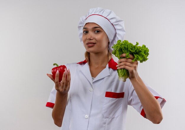 Cocinera joven complacida con uniforme de chef sosteniendo ensalada y pimienta en la pared blanca aislada