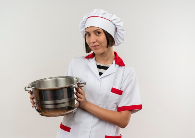 Cocinera joven complacida en uniforme de chef sosteniendo la olla mirando al frente aislado en la pared blanca