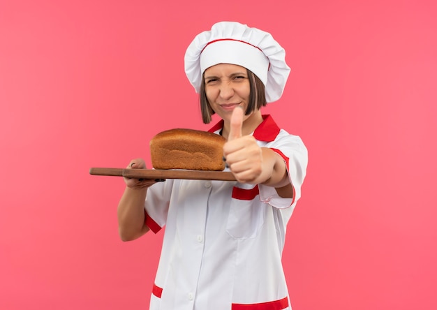 Cocinera joven complacida en uniforme de chef sosteniendo una tabla de cortar con pan y mostrando el pulgar hacia arriba aislado sobre fondo rosa con espacio de copia