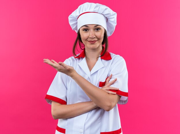 Cocinera joven complacida vistiendo puntos de uniforme de chef con la mano al lado aislado sobre fondo rosa con espacio de copia