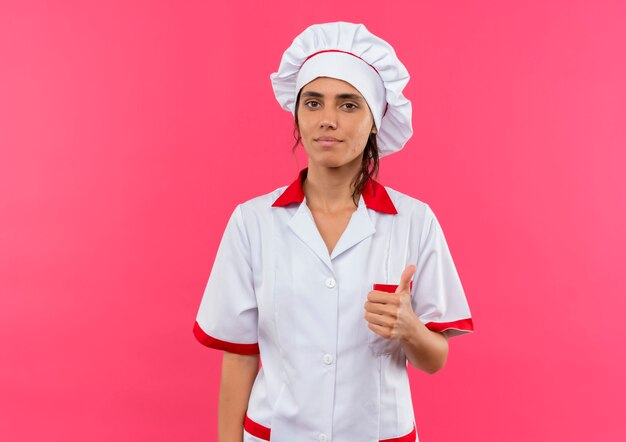 Cocinera joven complacida vistiendo uniforme de chef con el pulgar hacia arriba en la pared rosa aislada con espacio de copia