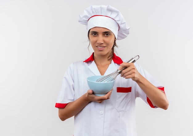 Cocinera joven complacida vistiendo uniforme de chef sosteniendo un batidor y un tazón con espacio de copia