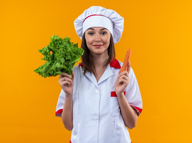 Cocinera joven complacida vistiendo uniforme de chef sosteniendo ensalada con zanahoria aislado en pared naranja