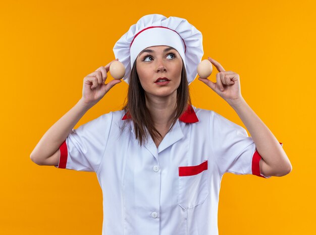 Cocinera joven complacida vistiendo uniforme de chef sosteniendo huevos alrededor de las orejas aislado en la pared naranja