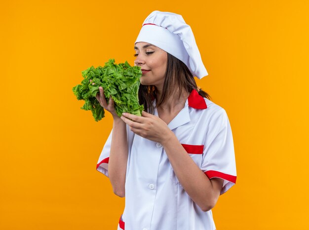 Cocinera joven complacida vistiendo uniforme de chef sosteniendo y oliendo ensalada aislada en pared naranja