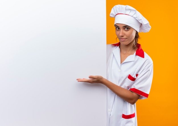 Cocinera joven complacida vistiendo uniforme de chef sosteniendo y puntos con mano pared blanca en pared amarilla aislada con espacio de copia