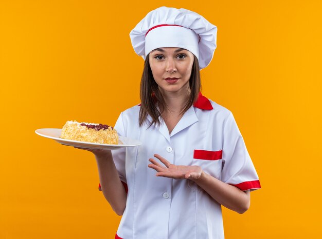 Cocinera joven complacida vistiendo uniforme de chef sosteniendo y puntos con la mano en el pastel en un plato aislado sobre fondo naranja