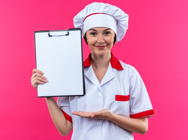 Cocinera joven complacida vistiendo uniforme de chef sosteniendo y puntos con la mano en el portapapeles aislado sobre fondo de color rosa