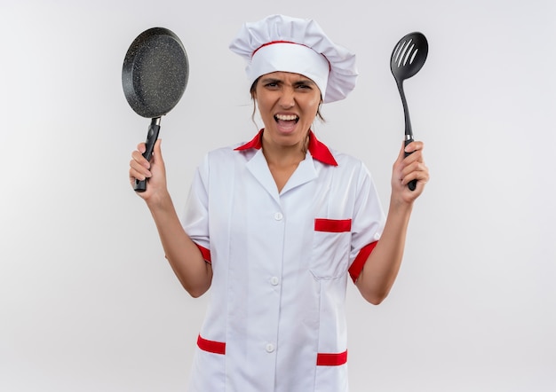 Cocinera joven enojada con uniforme de chef sosteniendo una sartén y una espátula en la pared blanca aislada con espacio de copia