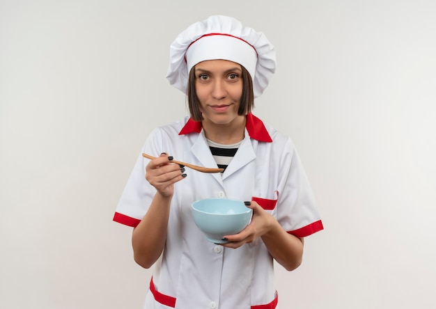 Cocinera joven feliz en uniforme de chef sosteniendo una cuchara y un tazón aislado en la pared blanca