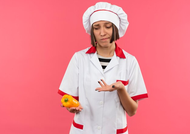 Cocinera joven pensativa en uniforme de chef sosteniendo mirando y apuntando con la mano a la pimienta aislada en la pared rosa