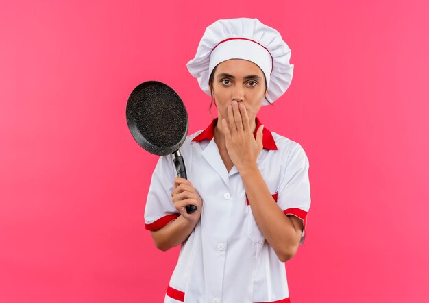 Cocinera joven sorprendida vistiendo uniforme de chef sosteniendo una sartén y la boca cubierta con la mano con espacio de copia