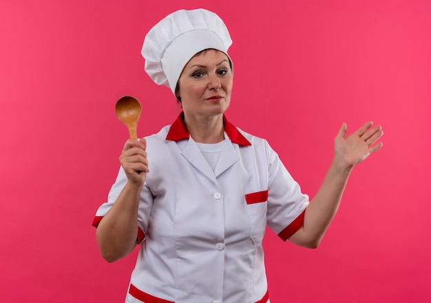 Cocinera de mediana edad en uniforme de chef sosteniendo una cuchara con la mano extendida en la pared rosa aislada