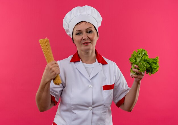 Cocinera de mediana edad en uniforme de chef sosteniendo ensalada y espaguetis en pared rosa aislada