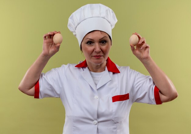 Cocinera de mediana edad en uniforme de chef sosteniendo huevos en la pared verde aislada