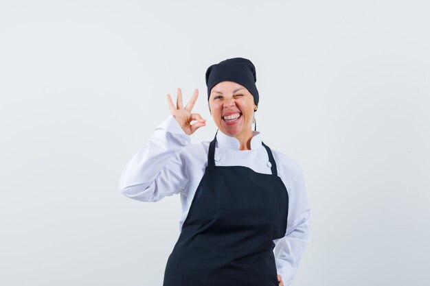 Cocinera mostrando gesto ok en uniforme, delantal y mirando dichoso. vista frontal.