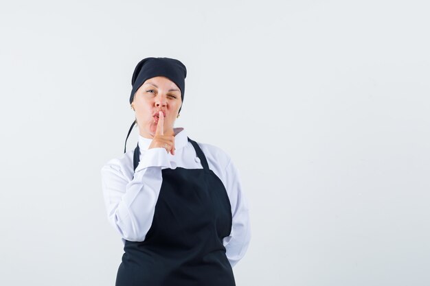 Cocinera mostrando gesto de silencio en uniforme, delantal y mirando confiado. vista frontal.