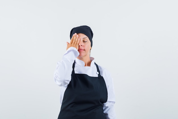 Cocinera sosteniendo la mano en el ojo en uniforme, delantal y mirando pacífica, vista frontal.