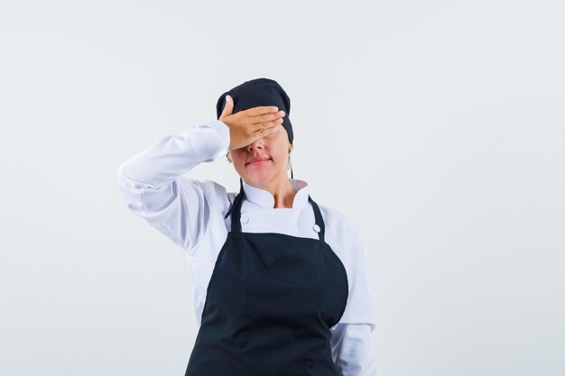 Cocinera sosteniendo la mano en los ojos en uniforme, delantal y mirando avergonzado, vista frontal.