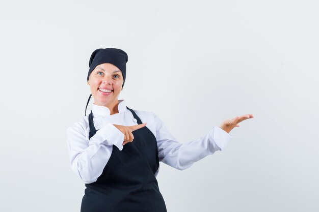 Cocinera en uniforme, delantal apuntando a su palma extendida a un lado y mirando confiada, vista frontal.