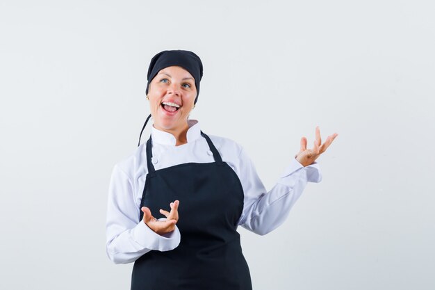 Cocinera en uniforme, delantal levantando las manos y mirando feliz, vista frontal.