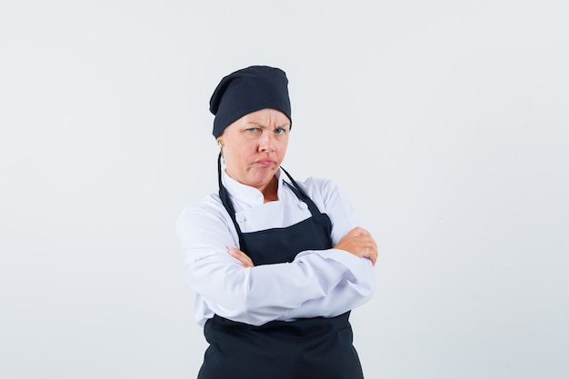 Cocinera en uniforme, delantal de pie con los brazos cruzados y mirando ofendido, vista frontal.