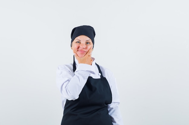 Cocinera en uniforme, delantal sosteniendo su barbilla y mirando vacilante, vista frontal.