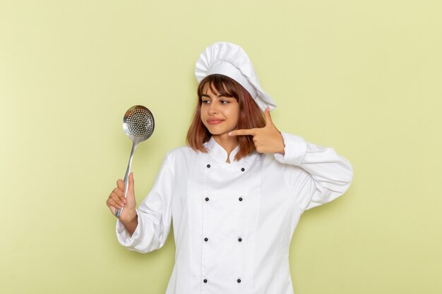 Cocinera de vista frontal en traje de cocinero blanco sosteniendo una cuchara de plata grande sobre una superficie verde claro