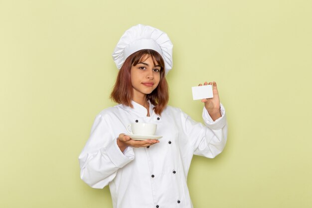 Cocinera vista frontal en traje de cocinero blanco sosteniendo una taza de té y una tarjeta en la superficie verde