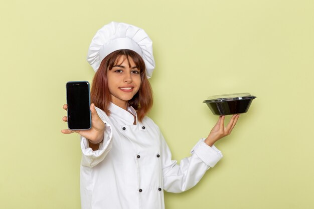 Cocinera de vista frontal en traje de cocinero blanco sosteniendo teléfono y cuenco negro sobre superficie verde