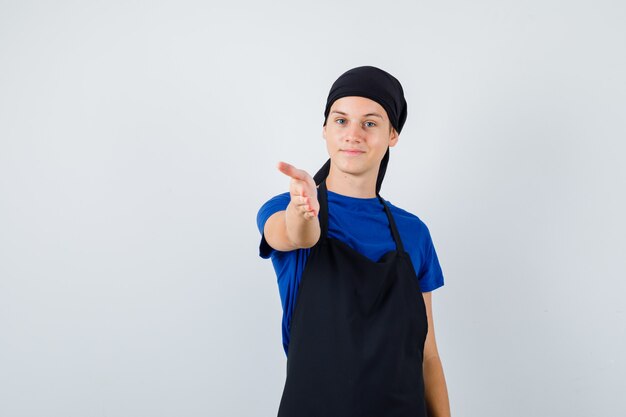 Cocinero adolescente joven que ofrece apretón de manos como saludo en camiseta, delantal y mirando alegre. vista frontal.