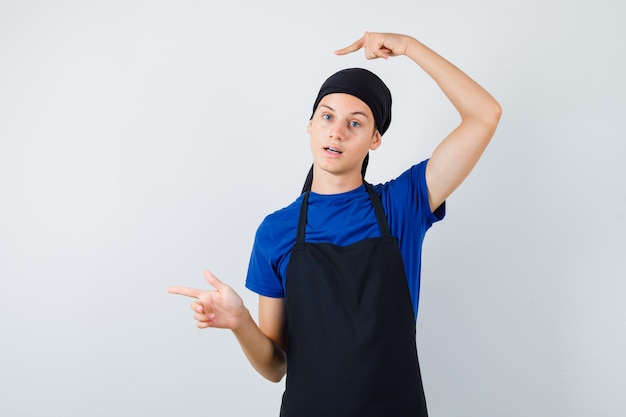 Cocinero adolescente masculino apuntando hacia el lado izquierdo en camiseta, delantal y mirando sorprendido, vista frontal.