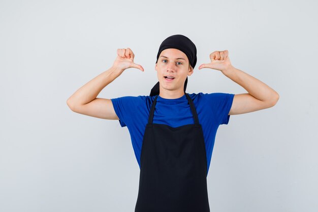 Cocinero adolescente masculino apuntando a sí mismo con los pulgares en camiseta, delantal y mirando confiado. vista frontal.
