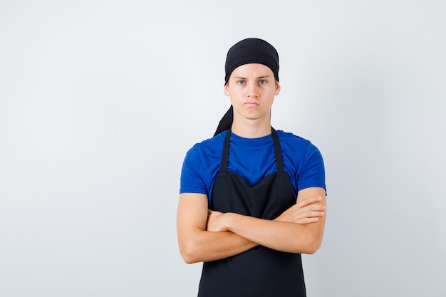 Cocinero adolescente masculino en camiseta, delantal con las manos cruzadas y mirando disgustado, vista frontal.