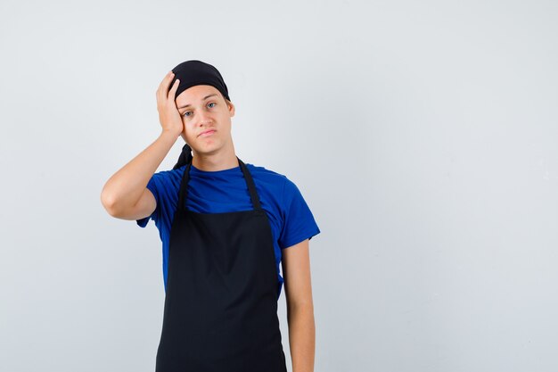 Cocinero adolescente masculino en camiseta, delantal manteniendo la mano en la cabeza y mirando arrepentido, vista frontal.