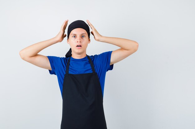 Cocinero adolescente masculino con las manos cerca de la cabeza, abriendo la boca en camiseta, delantal y mirando horrorizado, vista frontal.