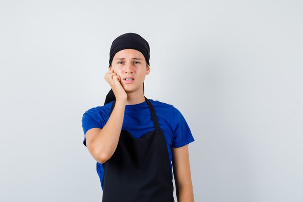 Cocinero adolescente masculino manteniendo la mano en la mejilla en camiseta, delantal y mirando disgustado. vista frontal.