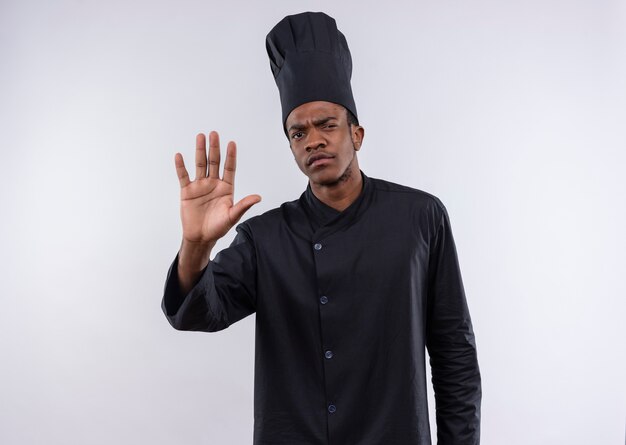 Cocinero afroamericano confiado joven en gestos uniformes del cocinero dejar de firmar la mano aislada sobre fondo blanco con espacio de copia