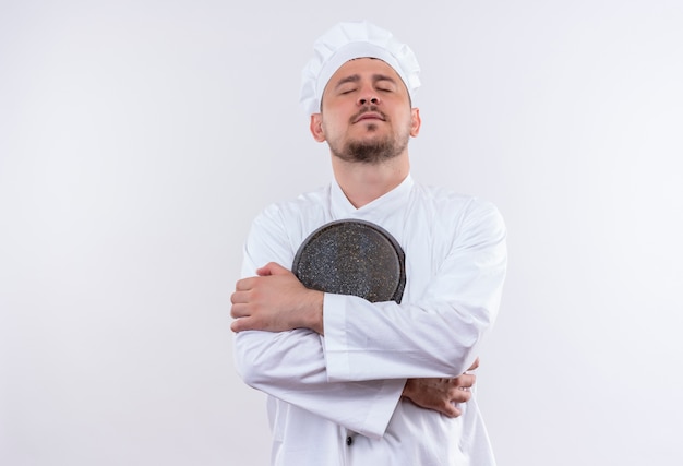 Cocinero apuesto joven pacífico en uniforme del cocinero que sostiene la sartén con los ojos cerrados en el espacio en blanco aislado