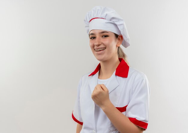 Cocinero bastante joven alegre en uniforme del cocinero con los apoyos dentales que aprietan el puño aislado en la pared blanca con el espacio de la copia