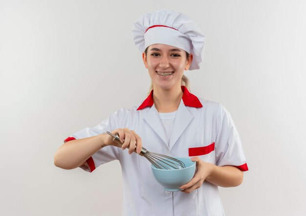 Cocinero bastante joven alegre en uniforme del cocinero con los apoyos dentales que sostienen el batidor y el cuenco aislado en el espacio blanco
