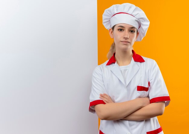 Cocinero bastante joven confiado en uniforme del cocinero que se coloca delante de la pared blanca con la postura cerrada aislada en la pared blanca