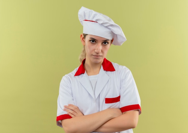 Cocinero bastante joven confiado en el uniforme del cocinero que se coloca con la postura cerrada aislada en la pared verde